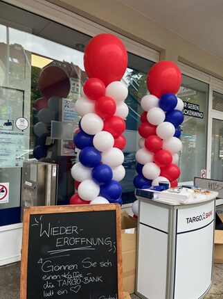 Zwei Säulen aus roten, weißen und blauen Ballons stehen vor einer Bankfiliale. Ein Schild mit der Aufschrift 'WIEDERERÖFFNUNG' und einer Einladung zur TARGO-BANK ist sichtbar.