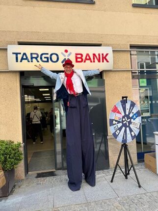Eine Person auf Stelzen steht vor der TARGOBANK. Sie trägt einen roten Hut, eine rote Krawatte und eine weiße Jacke. Neben ihr steht ein Glücksrad.