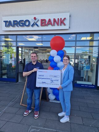Eine Person hält einen großen Scheck vor der TARGO BANK, während eine andere Person daneben steht. Im Hintergrund sind bunte Luftballons in Blau, Weiß und Rot, und das Bankgebäude ist klar sichtbar.