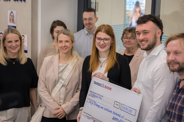 Eine Gruppe von Menschen steht in einer Bankfiliale. Im Vordergrund hält eine Frau mit Brille einen großen Scheck von TARGO BANK über 6000 Euro. Um sie herum stehen mehrere Personen, die lächeln und zuschauen. Die Atmosphäre ist freundlich und festlich.