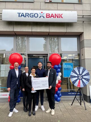 Fünf Personen stehen vor einer TARGO BANK-Filiale, die mit roten, weißen und blauen Luftballons dekoriert ist. Zwei Männer und eine Frau halten einen großen Scheck, während die anderen beiden Personen daneben stehen. Hinter ihnen sind die Fenster der Bank sichtbar, und ein Glücksrad ist ebenfalls im Bild zu sehen.
