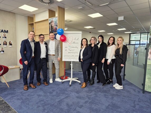 Eine Gruppe von acht Personen posiert in einem modernen Büro. Sie stehen vor einem Flipchart mit der Aufschrift 'Herzlich Willkommen zur 7. Geburtstag des ...'. Im Hintergrund sind helle Wände und Büromöbel zu sehen. Einige Personen tragen formelle Kleidung, während andere leger gekleidet sind. Rote und blaue Luftballons sind ebenfalls sichtbar.