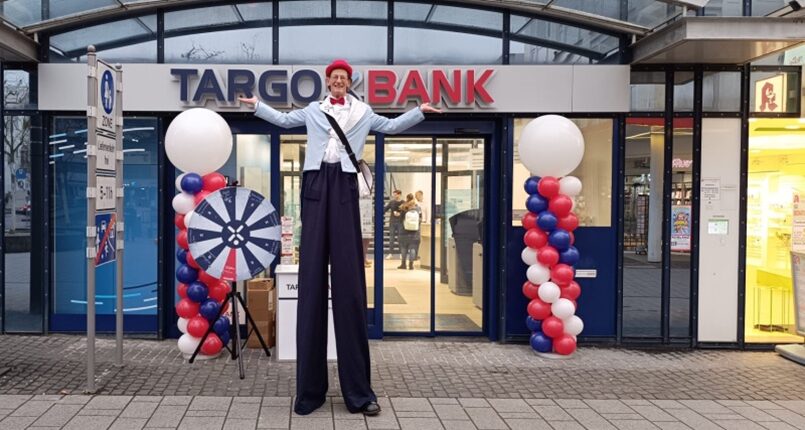 Vor einer Targobank-Filiale steht eine Person auf Stelzen. Die Person trägt eine weiße Bluse, eine rote Mütze und schwarze Hosen. Hinter ihr sind große, bunte Luftballons in Rot, Weiß und Blau aufgereiht. Links von der Person steht ein großes Dartboard, das ebenfalls rot und blau gestaltet ist. Der Eingang zur Bank ist gut erkennbar, mit mehreren Personen im Hintergrund.