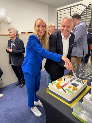 Zwei Personen schneiden zusammen eine Torte in einem modernen Raum. Die Frau trägt einen blauen Anzug und lächelt. Der Mann, in einem schwarzen Anzug, steht neben ihr. Im Hintergrund sind weitere Personen zu sehen. Auf dem Tisch liegt eine Torte mit der Aufschrift 'Happy Birthday' und essbaren Dekorationen.