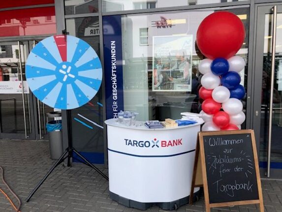 Ein Empfangstisch der TARGOBANK steht vor einem Eingang. Auf dem Tisch liegen Broschüren. Daneben steht ein Glücksrad mit blauer Farbe. Ein großes rotes Ballonarrangement ist ebenfalls sichtbar. Eine Tafel begrüßt die Gäste zur Jubiläumsfeier der TARGOBANK.