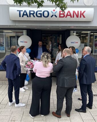 Vor der TARGO BANK stehen mehrere Personen, darunter Männer in Anzügen und Frauen in eleganter Kleidung. Eine Frau hält einen Blumenstrauß und es gibt Luftballons in den Farben Blau und Weiß. Die Szene wirkt festlich und es scheint, als ob eine Eröffnungsfeier oder ein Event stattfindet.