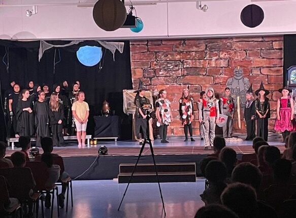 Eine Theateraufführung mit einer Gruppe von Darstellern auf der Bühne. Sie stehen in verschiedenen Kostümen, darunter schwarze Kleidung, farbenfrohe Outfits und historische Kostüme. Im Hintergrund sind große Steine und ein Fenster sichtbar. Das Publikum sitzt im Dunkeln und beobachtet die Aufführung, während einige Darsteller in den Vordergrund treten.