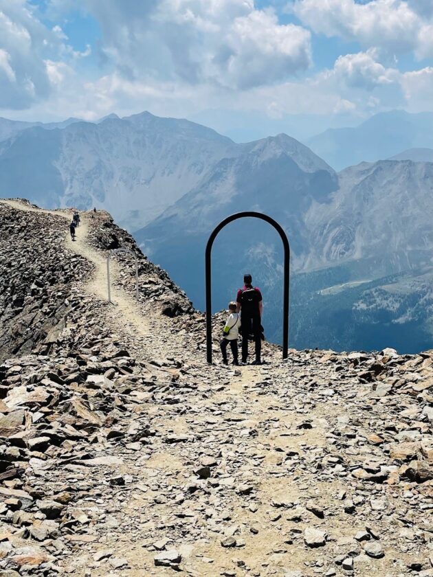 Eine Wanderung auf einem schmalen Pfad, der sich entlang eines Bergkamms schlängelt. Im Vordergrund stehen ein Erwachsener und ein Kind neben einem großen, schwarzen, rechteckigen Rahmen, der in die Landschaft hineinragt. Im Hintergrund sind majestätische Berge und ein blauer Himmel mit Wolken sichtbar. Der Boden ist steinig und uneben.