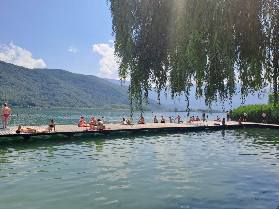 Ein See mit vielen Menschen, die auf einem Holzsteg sitzen und sich sonnen. Einige schwimmen im Wasser, während andere auf Handtüchern liegen. Im Hintergrund sind grüne Berge und ein blauer Himmel mit wenigen Wolken sichtbar. Ein Weidenbaum hängt über den Steg.