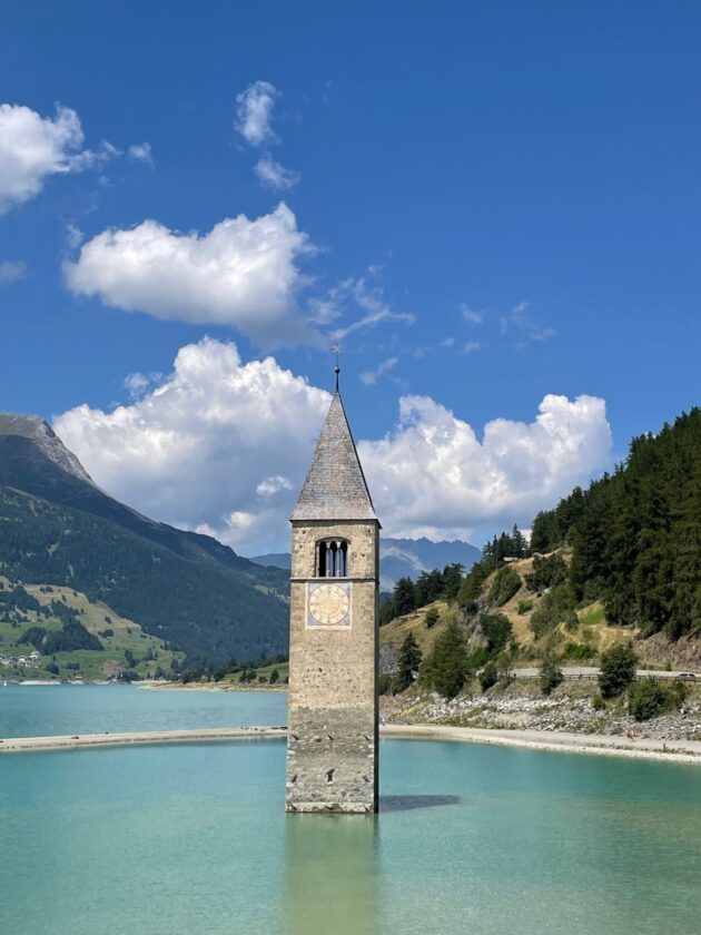 Ein alter Glockenturm mit einem dreieckigen Dach steht in einem klaren, türkisfarbenen See. Im Hintergrund sind grüne Hügel und Berge sichtbar, während der Himmel blau mit einigen weißen Wolken ist.