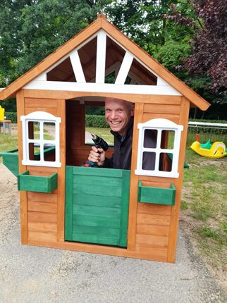 Ein Mann sitzt in einem kleinen Holzhaus mit einem grünen Tor und weißen Fenstern. Er hält eine Bohrmaschine in der Hand und lächelt. Das Haus steht auf einem Spielplatz mit grünen Bäumen im Hintergrund.