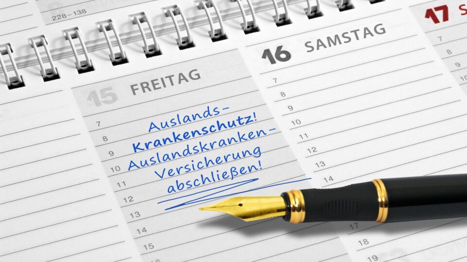 Ein Kalenderblatt zeigt den Freitag, den 15. mit den Notizen "Auslands-Krankenschutz!" und "Auslands-Kranken-Versicherung abschließen!". Ein Füllfederhalter liegt am unteren Rand.