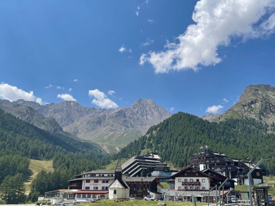 Ein malerisches Bergdorf mit mehreren Gebäuden, darunter ein modernes Hotel und eine traditionelle Kirche. Im Hintergrund erstrecken sich hohe Berge unter einem klaren blauen Himmel mit einigen Wolken. Die Landschaft ist grün bewaldet, und eine Wiese ist im Vordergrund sichtbar.
