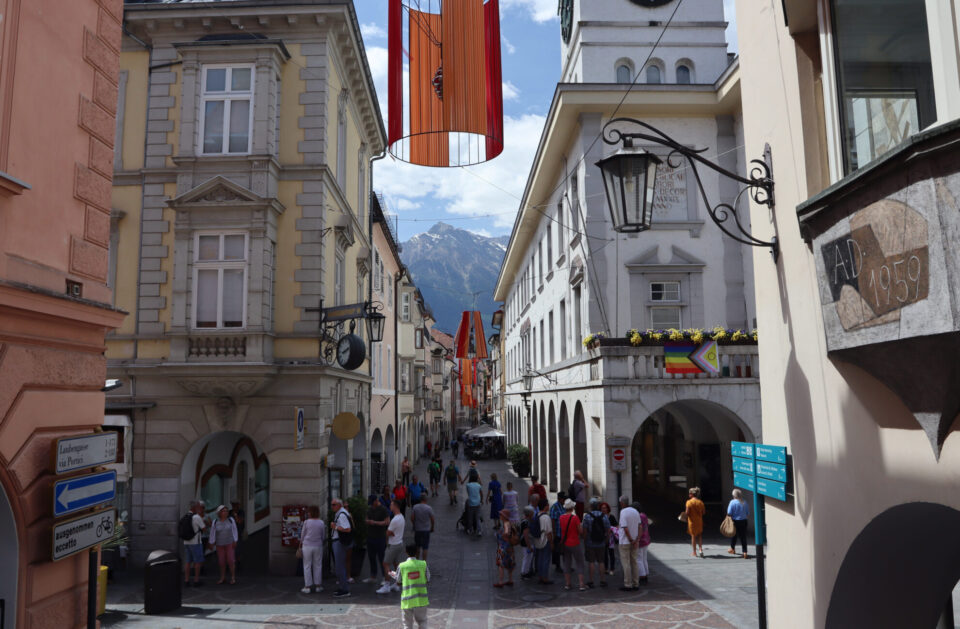 Eine belebte Straße mit historischen Gebäuden, an denen orangefarbene Dekoration hängt. Menschen stehen und gehen, während die Berglandschaft im Hintergrund sichtbar ist. Eine Uhr und Straßenschilder sind ebenfalls zu sehen.