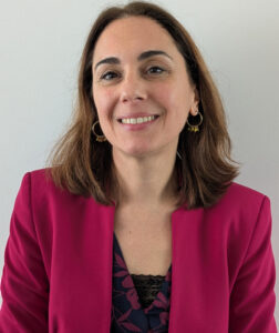 Eine Frau mit langen, braunen Haaren lächelt in die Kamera. Sie trägt einen pinken Blazer über einem dunkelblauen Oberteil mit floralem Muster und Spitzenkragen. Ihr Look wird durch goldene Ohrringe ergänzt. Der Hintergrund ist schlicht und weiß.