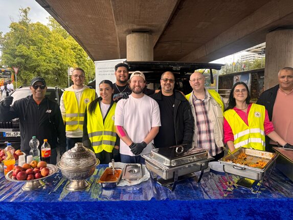 Eine Gruppe von neun Personen steht an einem Tisch unter einer Brücke. Alle tragen reflektierende Westen. Vor ihnen stehen verschiedene Gerichte in Warmhaltebehältern sowie Obst. Zwei Personen halten eine Getränkeflasche. Im Hintergrund sind Bäume und ein Fahrzeug sichtbar.