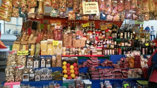 Ein belebter Marktstand mit einer Vielzahl von Lebensmitteln. Oben hängen bunte Teigtaschen und Nudeln. Darunter sind Flaschen mit Öl und Spirituosen, verschiedene Marmeladen und Trockenfrüchte. Auf dem Tisch sind bunte Äpfel und verschiedene Snacks ausgestellt. Verkaufsangebote sind gut sichtbar, darunter "3 x Nudeln, 1 x gratis".