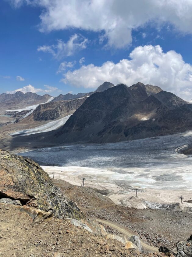 Eine beeindruckende Berglandschaft mit schroffen Gipfeln, die unter einem blauen Himmel mit weißen Wolken liegen. Im Vordergrund sind steinige Hänge und ein Gletscher sichtbar, während im Hintergrund weitere Berge zu sehen sind. Eine Skipiste oder Seilbahnen sind am unteren Rand des Bildes angedeutet, die den Zugang zur schneebedeckten Fläche ermöglichen.