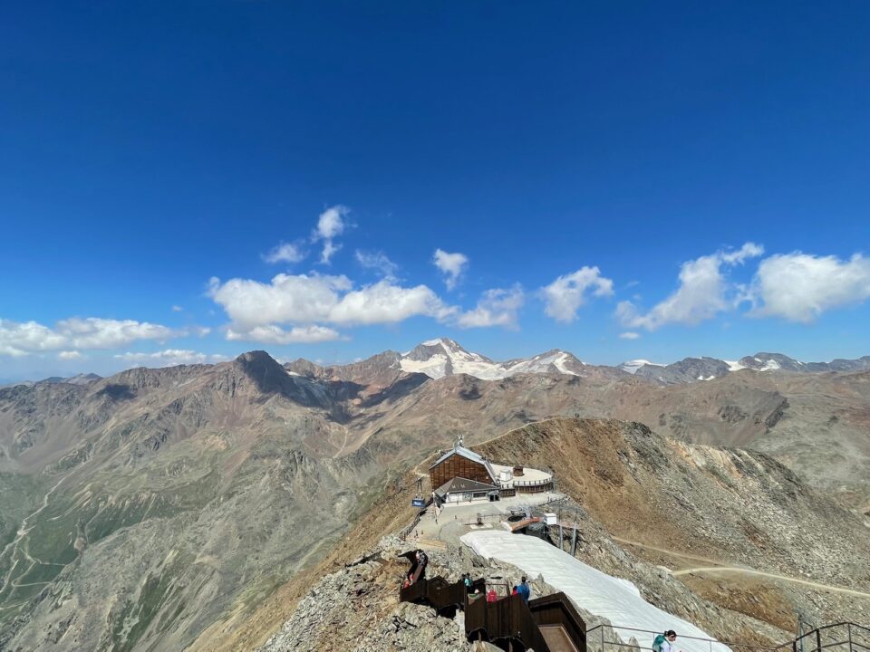 Eine beeindruckende Berglandschaft unter einem klaren blauen Himmel mit ein paar Wolken. Im Vordergrund ist eine Berghütte sichtbar, umgeben von felsigem Terrain und alpinen Gipfeln. Im Hintergrund erheben sich schneebedeckte Berge, und einige Wanderer sind auf dem Weg. Die Landschaft zeigt eine Mischung aus steilen Hängen und sanften Tälern.