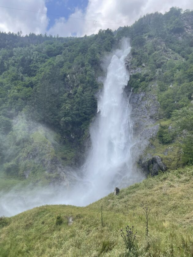 Ein Wasserfall strömt über eine felsige Klippe in eine grüne Landschaft. Der Wasserfall erzeugt Gischt und Wasserdampf, umgeben von üppigem Wald und Hügeln. Der Himmel ist teilweise bewölkt mit blauen Abschnitten.
