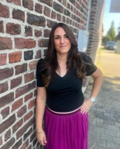 Eine Frau mit langen, lockigen Haaren steht an einer Backsteinmauer. Sie trägt ein schwarzes T-Shirt und einen langen, lila Rock. Ihre linke Hand ist auf ihrer Hüfte, während sie lächelt. Im Hintergrund sind unscharfe Gebäude und ein gepflasterter Weg sichtbar.