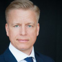 Ein Mann mit blonden, kurzen Haaren und blauen Augen. Er trägt einen dunklen Anzug mit einer hellen Krawatte. Der Hintergrund ist dunkel.