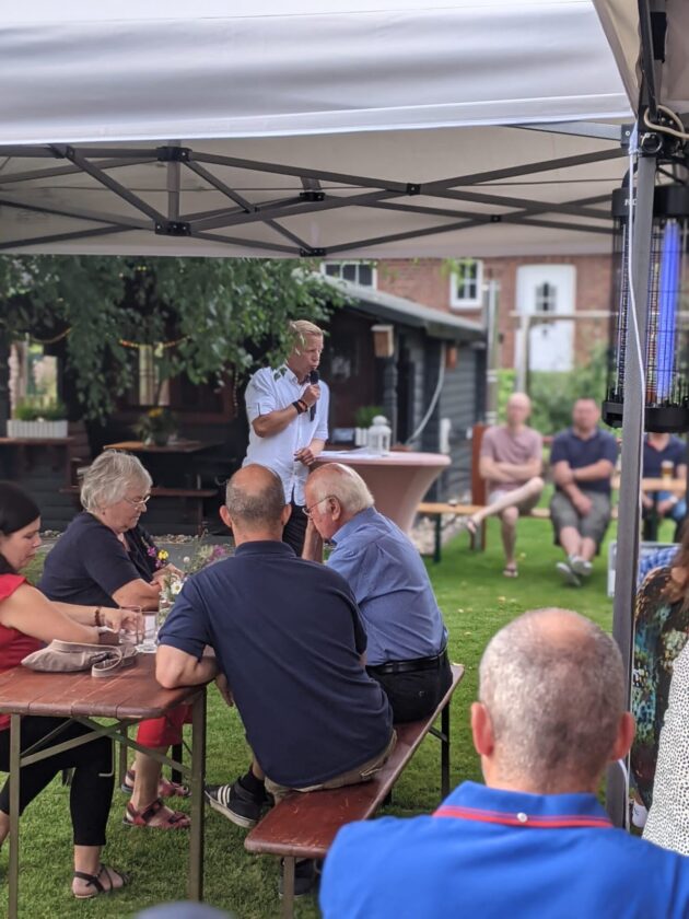Eine Outdoor-Veranstaltung unter einem Zelt mit Gästen, die an Tischen sitzen. Ein Mann in einem weißen Hemd spricht mit einem Mikrofon, während einige Gäste ihm zuhören. Ältere Frauen und Männer sind an den Tischen versammelt, es gibt Blumen in der Mitte des Tisches. Im Hintergrund sind Bäume und ein Holzgebäude sichtbar.