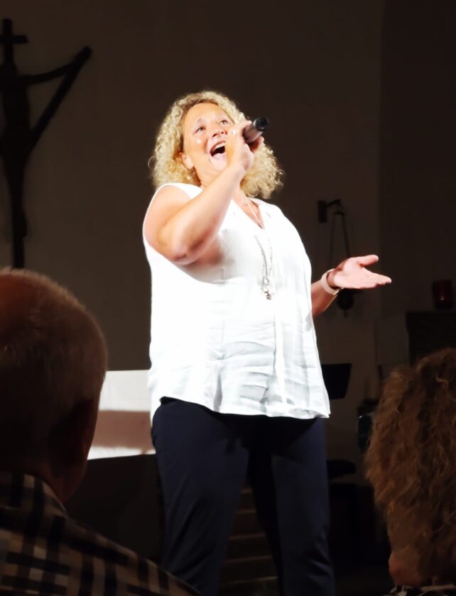 Eine Frau mit lockigem, blondem Haar singt leidenschaftlich in einem Raum. Sie trägt ein weißes Oberteil und blaue Hosen, hält ein Mikrofon in der Hand und hat einen Ausdruck der Freude im Gesicht. Im Hintergrund ist eine schlichte Dekoration zu sehen, sowie eine dunkle Silhouette eines Kreuzes an der Wand.