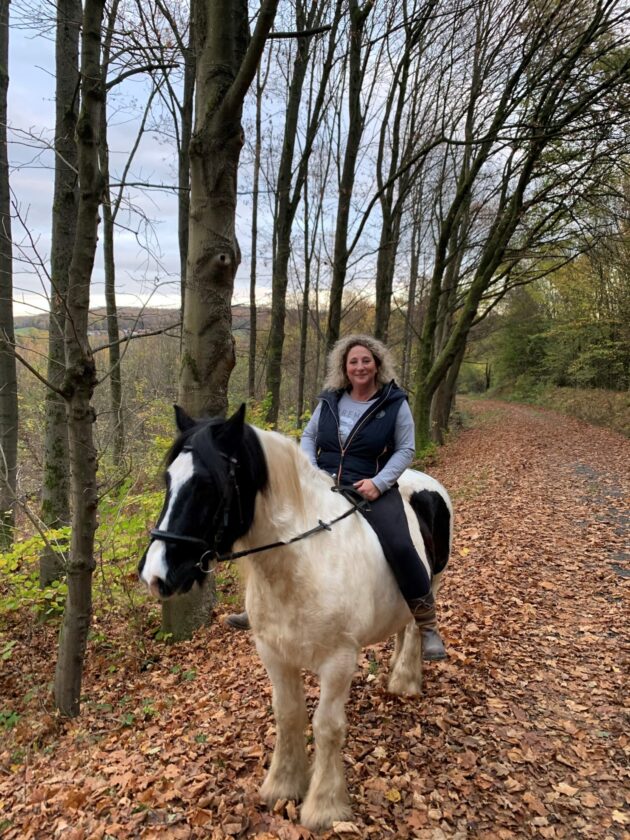 Eine Frau mit lockigem, blondem Haar reitet auf einem schwarz-weißen Pferd. Sie trägt eine graue Jacke und sitzt auf einem Weg, umgeben von Bäumen und buntem Laub im Herbst.