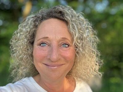 Eine Frau mit lockigem, blondem Haar und blauen Augen lächelt in die Kamera. Der Hintergrund ist grün und verschwommen.