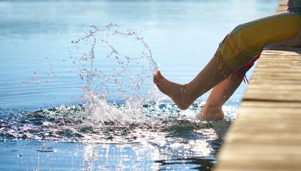 Ein Fuß spritzt Wasser, während er in einem ruhigen Gewässer hängt. Der Fuß befindet sich am Rand eines Holzdecks, umgeben von sanften Wellen und Sonnenlicht, das auf der Wasseroberfläche reflektiert.