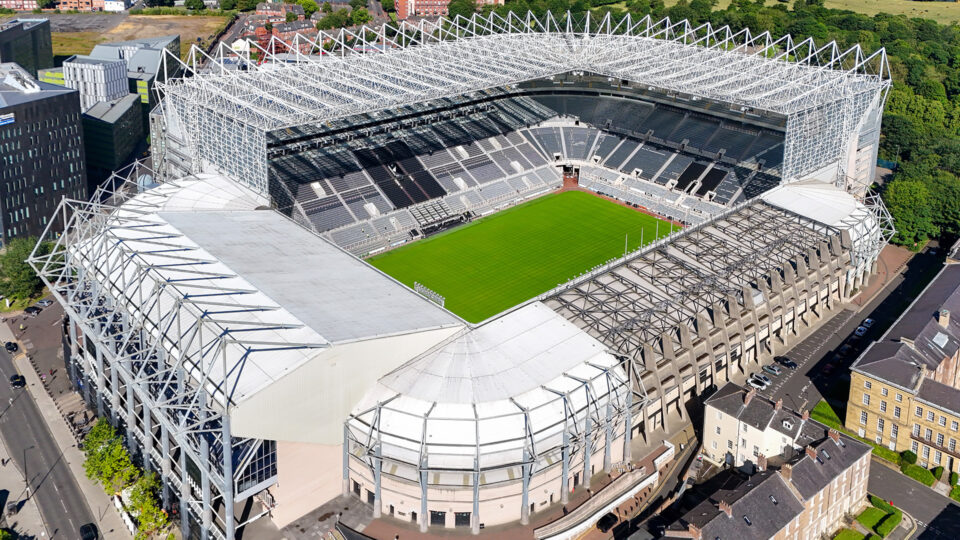 Eine Luftaufnahme eines großen Fußballstadions mit einem grünen Spielfeld in der Mitte. Die Tribünen sind leer und das Stadion hat ein modernes, offenes Design mit einem markanten Dach. Um das Stadion herum sind einige Gebäude und Straßen sichtbar.