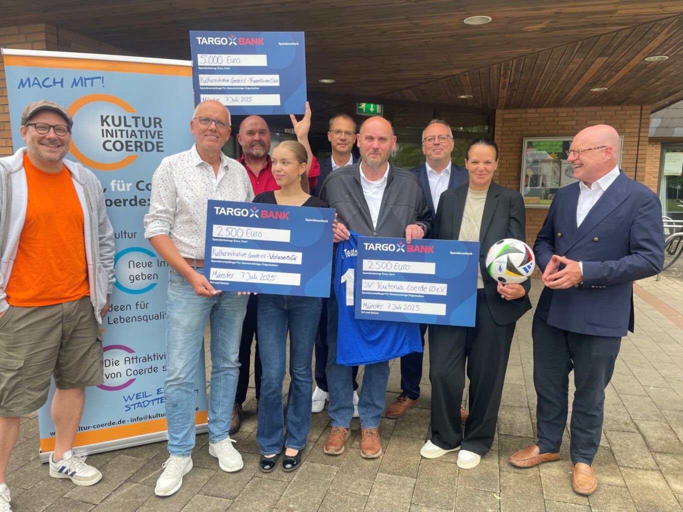Eine Gruppe von sechs Personen steht vor einem Banner der Kulturinitiative Coerde. Sie halten Schecks der TARGOBANK in Höhe von 5.000 Euro und zwei Schecks über 2.500 Euro. Die Personen lächeln, während sie die Schecks präsentieren. Im Hintergrund ist ein Gebäude sichtbar. Das Datum auf den Schecks ist der 7. Juli 2025.