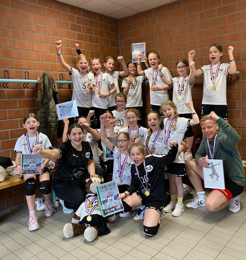 Eine Gruppe von Mädchen in Sportkleidung feiert in einem Umkleideraum. Sie tragen Medaillen und halten Urkunden in den Händen. Einige Mädchen haben die Arme erhoben und lächeln, während andere jubeln. Im Hintergrund sind hängende Kleiderhaken und eine Wand mit Ziegelsteinen sichtbar. Ein Trainer sitzt auf einer Bank und lächelt.