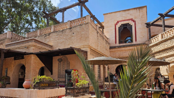 Eine Außenansicht eines traditionellen Gebäudes mit Steinmauern und einem offenen Café-Bereich. In der Mitte ist ein Tisch mit Sonnenschirmen, umgeben von Pflanzen. Im Hintergrund sind Fenster mit dekorativen Gitterelementen sichtbar. Menschen sitzen am Tisch und unterhalten sich, während ein großer Baum die Szene beschattet.