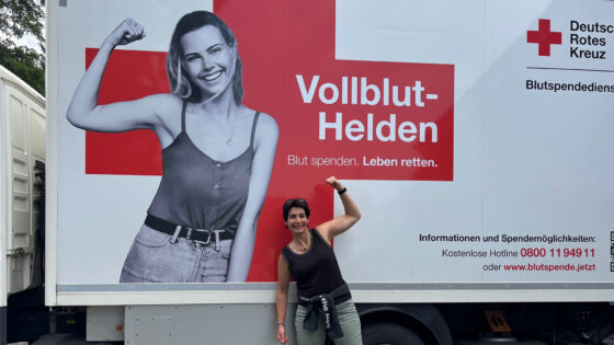 Ein Werbeanhänger des Deutschen Roten Kreuzes zeigt eine lächelnde Frau mit hochgestelltem Arm. Der Text "Vollblut-Helden" und "Blut spenden. Leben retten." ist sichtbar. Eine weitere Person steht daneben und posiert mit erhobenem Arm.