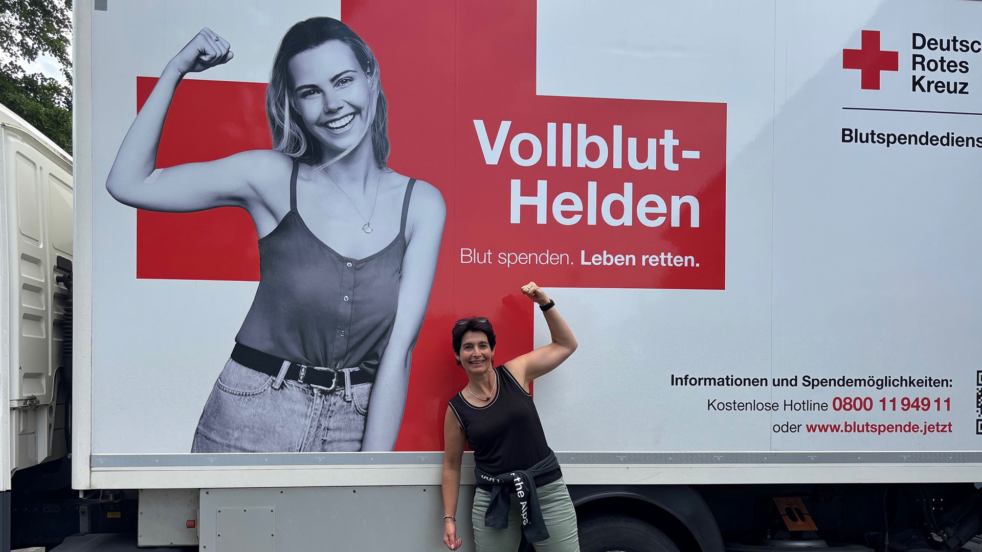 Ein Werbeanhänger des Deutschen Roten Kreuzes zeigt eine lächelnde Frau mit hochgestelltem Arm. Der Text "Vollblut-Helden" und "Blut spenden. Leben retten." ist sichtbar. Eine weitere Person steht daneben und posiert mit erhobenem Arm.