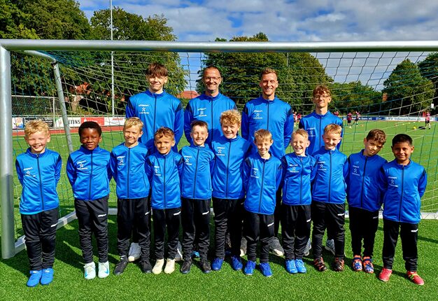 Eine Gruppe von Fußballspielern und Trainern steht vor einem Tor auf einem Sportplatz. Die Spieler tragen blaue Trainingsjacken und schwarze Hosen. Es gibt fünf Trainer und zehn Kinder in der Gruppe. Die Spieler lächeln und posieren für das Foto, während im Hintergrund andere Kinder Fußball spielen. Der Himmel ist klar mit einigen Wolken.