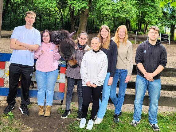 Eine Gruppe von sieben Menschen steht im Freien, umgeben von Bäumen. Sie posieren lächelnd für ein Foto. In der Mitte steht ein Pferd, das sich den Personen nähert. Die Menschen tragen unterschiedliche Freizeitkleidung. Es gibt eine Mischung aus langen und kurzen Hosen. Der Hintergrund zeigt eine Pferdeweide mit Zäunen.
