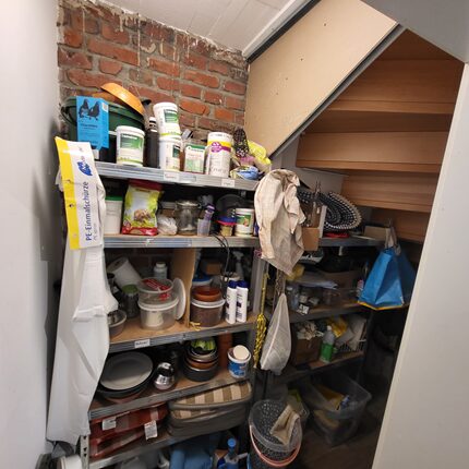 Ein Blick auf einen kleinen Abstellraum unter einer Treppe. Regale sind voll mit verschiedenen Behältern, Gläsern, Reinigungsmitteln und Küchenutensilien. Die Wände sind aus Ziegeln, und die Treppe führt nach oben. Auf dem Boden liegen einige weitere Gegenstände und Tüten.