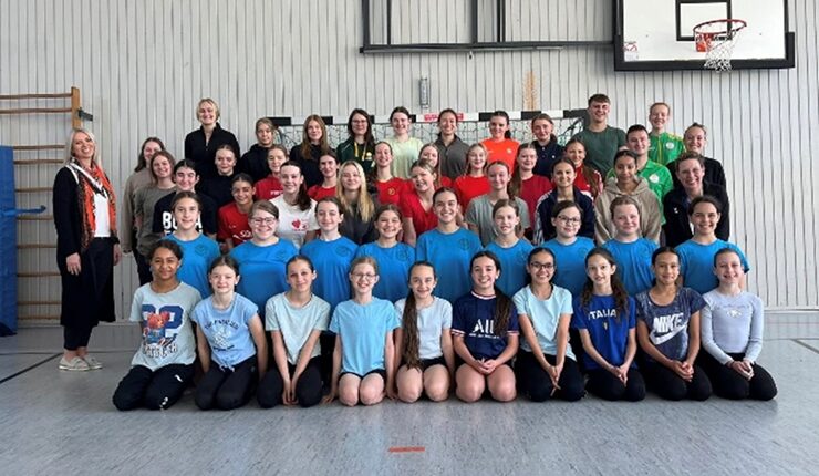 Eine große Gruppe von etwa 50 Mädchen und jungen Frauen steht in einer Sporthalle. Sie tragen verschiedene Sportbekleidungen, hauptsächlich T-Shirts und Shorts. Einige haben auch Sweatshirts an. Im Hintergrund ist ein Basketballkorb und eine Wand mit Holzgestellen sichtbar. Die Gruppe ist fröhlich und posiert für das Foto, während einige Erwachsene hinter ihnen stehen.