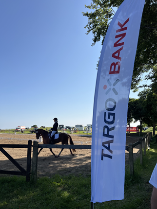 Ein Reiter in schwarzer Kleidung reitet auf einem braunen Pferd in einem Reitstall. Im Vordergrund steht ein großes Banner der TARGOBANK. Der Himmel ist blau und es ist sonnig.