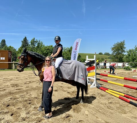 Eine Frau steht neben einem Pferd, während ein Reiter auf dem Pferd sitzt. Die Reiterin trägt einen Helm und eine schwarze Reithose. Die Frau hat blonde Haare und trägt ein schwarzes Outfit. Im Hintergrund sind Hindernisse für den Springreitwettbewerb zu sehen, und einige Personen bereiten sich vor. Der Himmel ist blau mit wenigen Wolken.