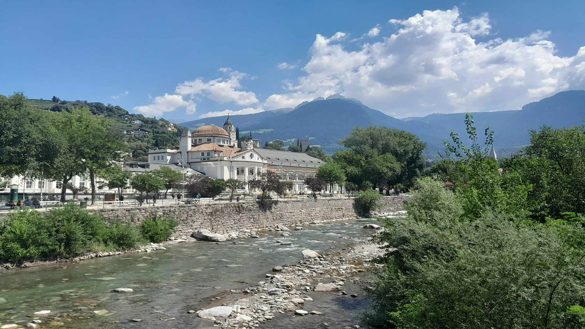 Ein malerischer Fluss fließt durch eine grüne Landschaft, umgeben von Bäumen und Pflanzen. Auf der linken Seite steht ein historisches Gebäude mit einer Kuppel und einem hohen Turm. Im Hintergrund erheben sich majestätische Berge unter einem blauen Himmel mit einigen Wolken. Spaziergänger sind entlang des Ufers zu sehen, was der Szene Leben verleiht.