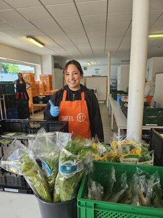 Eine junge Frau in einem orangefarbenen Schürze mit dem Logo 'TAFEL' steht in einem Raum mit vielen Kisten. Sie trägt blaue Handschuhe und lächelt, während sie Daumen hoch zeigt. Im Vordergrund sind grüne Kisten mit frisch verpacktem Gemüse.