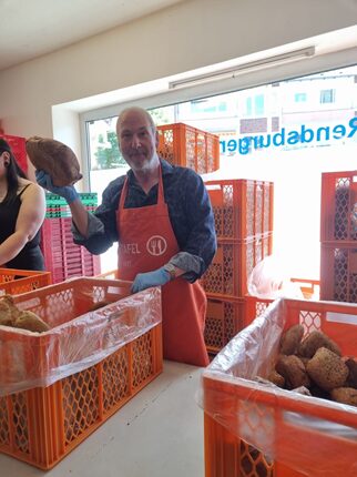 Ein Mann mit Glatze und lächelndem Gesicht trägt eine orangefarbene Schürze. Er steht in einem Raum mit orangefarbenen Kisten, die mit gebackenem Brot gefüllt sind. Der Mann hält ein großes Brot in der einen Hand und zeigt es stolz. Im Hintergrund sind weitere Kisten und eine Fensterscheibe zu sehen, auf der der Schriftzug 