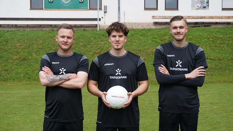 Drei Fußballspieler stehen auf einem Rasenplatz. Sie tragen schwarze Trikots mit dem Logo der TARGOBANK. Der mittlere Spieler hält einen Fußball in den Händen, während die anderen beiden Spieler auf beiden Seiten stehen. Im Hintergrund sind einige Fenster und eine Tribüne sichtbar.