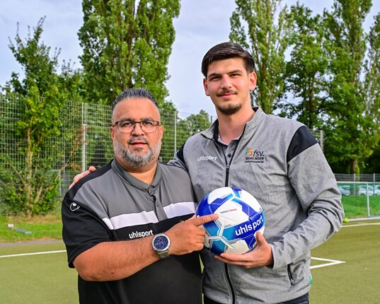 Zwei Männer stehen auf einem Sportplatz. Der Mann links trägt ein schwarzes und graues Trikot mit dem Logo von uhlsport und eine Brille. Er hat einen Bart und hält einen Fußball in der Hand. Der Mann rechts trägt ein graues Sportoberteil mit einer schwarzen Jacke und hat dunkles Haar. Beide lächeln in die Kamera und stehen inmitten von Bäumen und einem Fußballfeld.