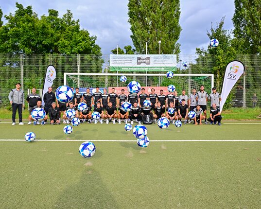 Eine Gruppe von Fußballspielern steht auf einem Fußballfeld vor einem Tor. Sie tragen einheitliche schwarze Trikots und posieren für ein Gruppenfoto. Im Vordergrund sind viele Fußbälle mit blauen und weißen Mustern zu sehen, die in der Luft schweben. Im Hintergrund sind Bäume und ein Banner sichtbar, das den Verein oder das Event bewirbt. Der Himmel ist teilweise bewölkt.
