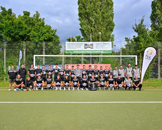 Eine Gruppe von Fußballspielern und Trainern steht auf einem Fußballfeld vor einem Tor. Sie tragen einheitliche schwarze Trikots und halten Fußbälle. Im Hintergrund sind grüne Bäume und ein Banner sichtbar. Die gesamte Gruppe ist auf einer Kunstrasenfläche versammelt.